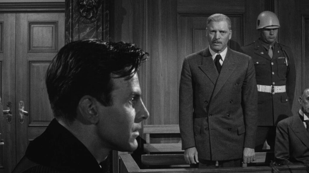 Im Vordergrund sieht man Maximilian Schell im Profil, dahinter steht Burt Lancester als Angeklagter, hinter ihm ein Soldat - Eine Szene aus "Das Urteil von Nürnberg" (1961)
