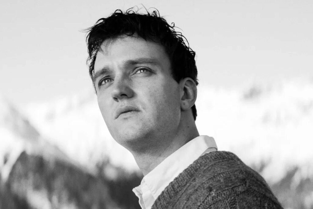Jan Bülow als Johannes Leinert in „Die Theorie von Allem“, in einem schwarz‑weißen Porträt vor einer verschneiten Berglandschaft. Er trägt einen grob gestrickten Pullover über einem Hemd und blickt leicht nach oben, während das helle Licht seine Gesichtszüge kontrastreich hervorhebt. | Media Monday #775