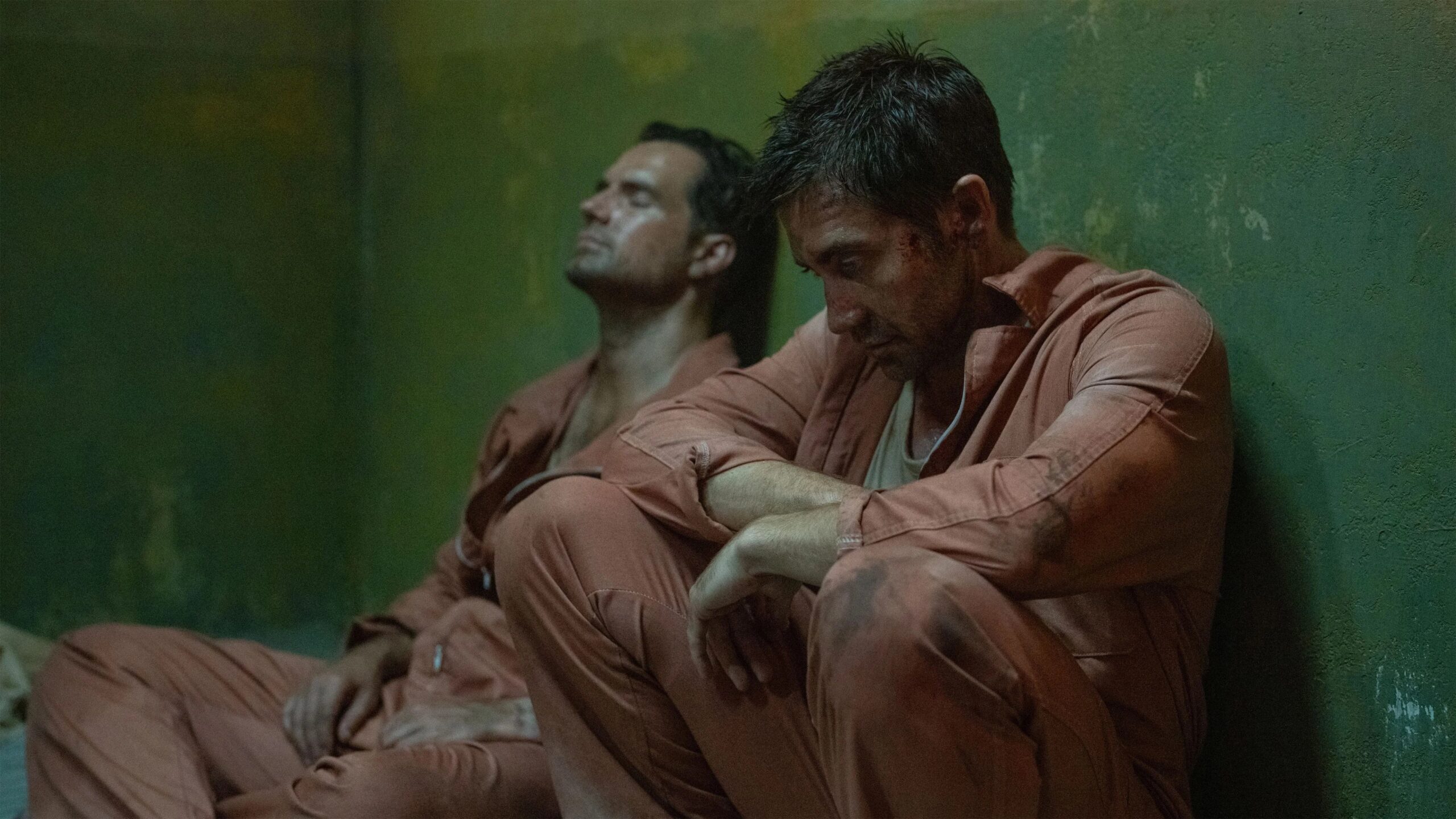 Bronco (Jake Gyllenhaal, vorne) sitzt erschöpft in einem engen, abgenutzten Raum in einem orangefarbenen Overall, während ein weiterer Gefangener neben ihm an der Wand lehnt. Beide wirken verschmutzt und ausgelaugt, die grünlich‑abgeblätterte Wand und das gedämpfte Licht unterstreichen die bedrückende, klaustrophobische Stimmung der Szene aus In the Grey.