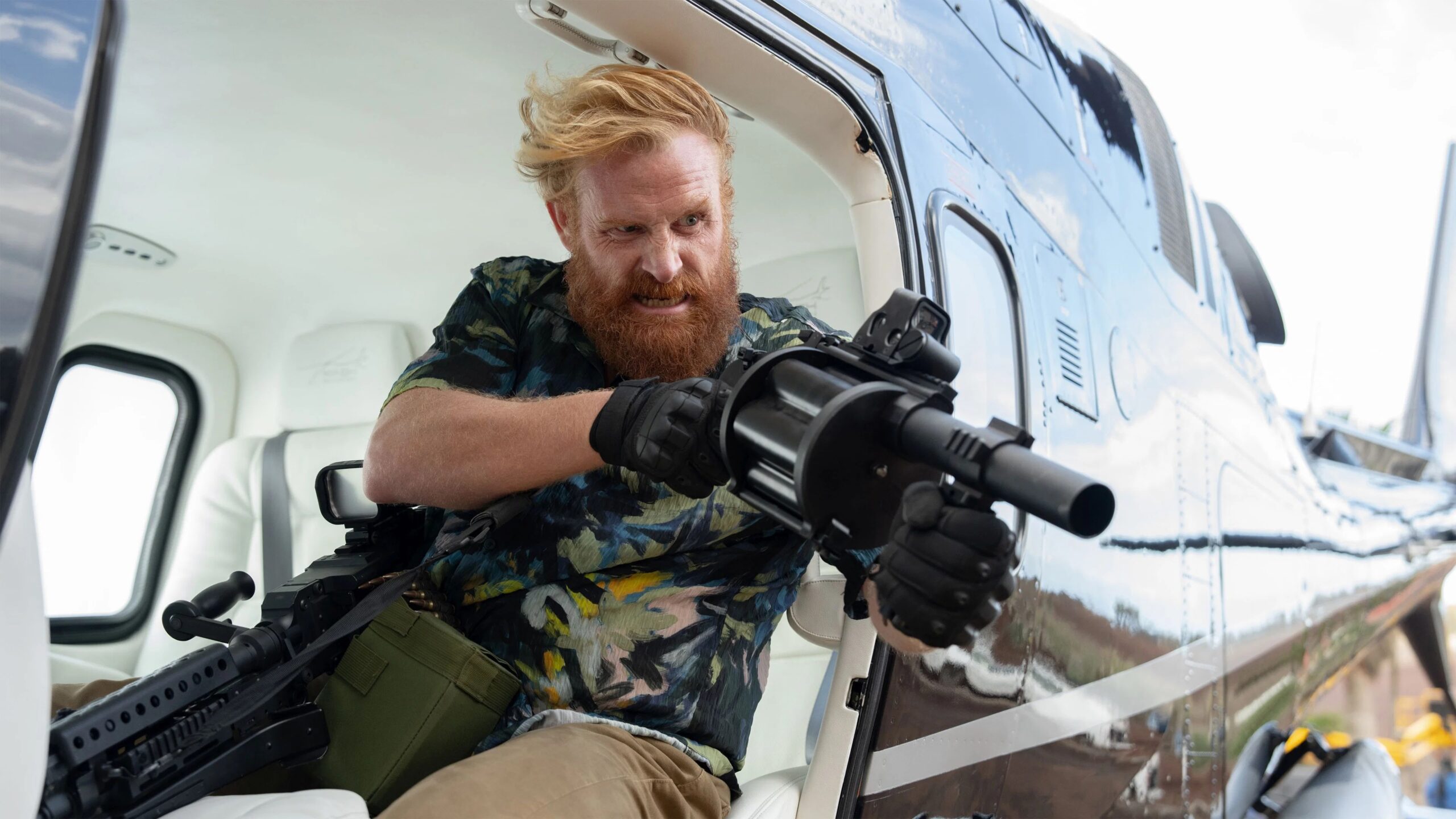 Kristofer Hivju im Film In the Grey sitzt im offenen Seiteneinstieg eines Helikopters, das Haar vom Wind zerzaust, und hält ein schweres, mehrläufiges Geschütz im Anschlag. Der metallisch glänzende Helikopter und die weißen Sitze bilden einen starken Kontrast zur dynamischen, actiongeladenen Haltung der Figur.