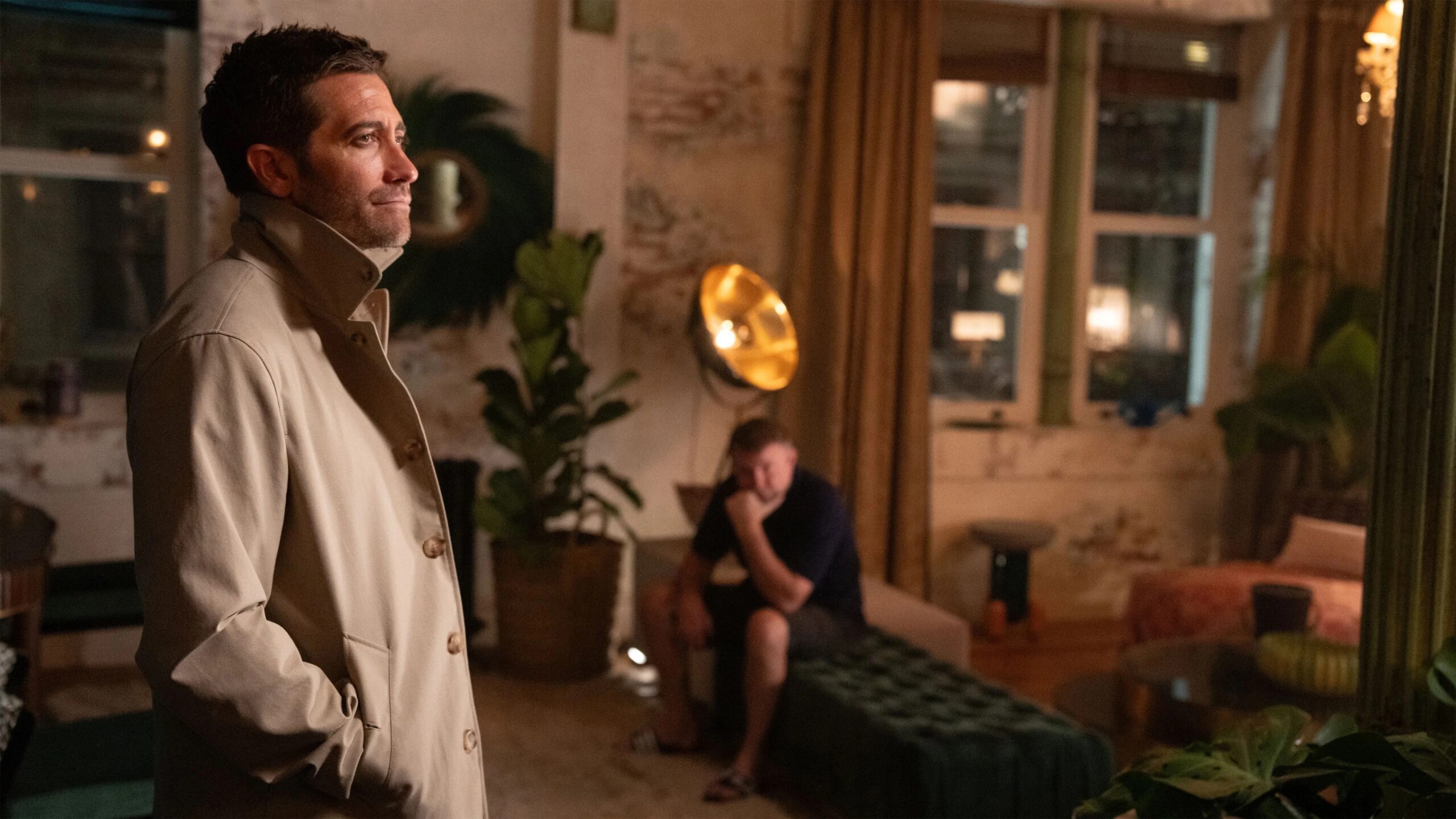 Jake Gyllenhaal im Film In the Grey steht in einem warm ausgeleuchteten, stilvollen Raum in einem beigefarbenen Mantel mit den Händen in den Taschen, während im Hintergrund eine zweite Person auf einem Sofa sitzt und nachdenklich den Kopf auf die Hand stützt. Große Fenster, Pflanzen und eine goldene Stehlampe erzeugen eine ruhige, intime Atmosphäre.
