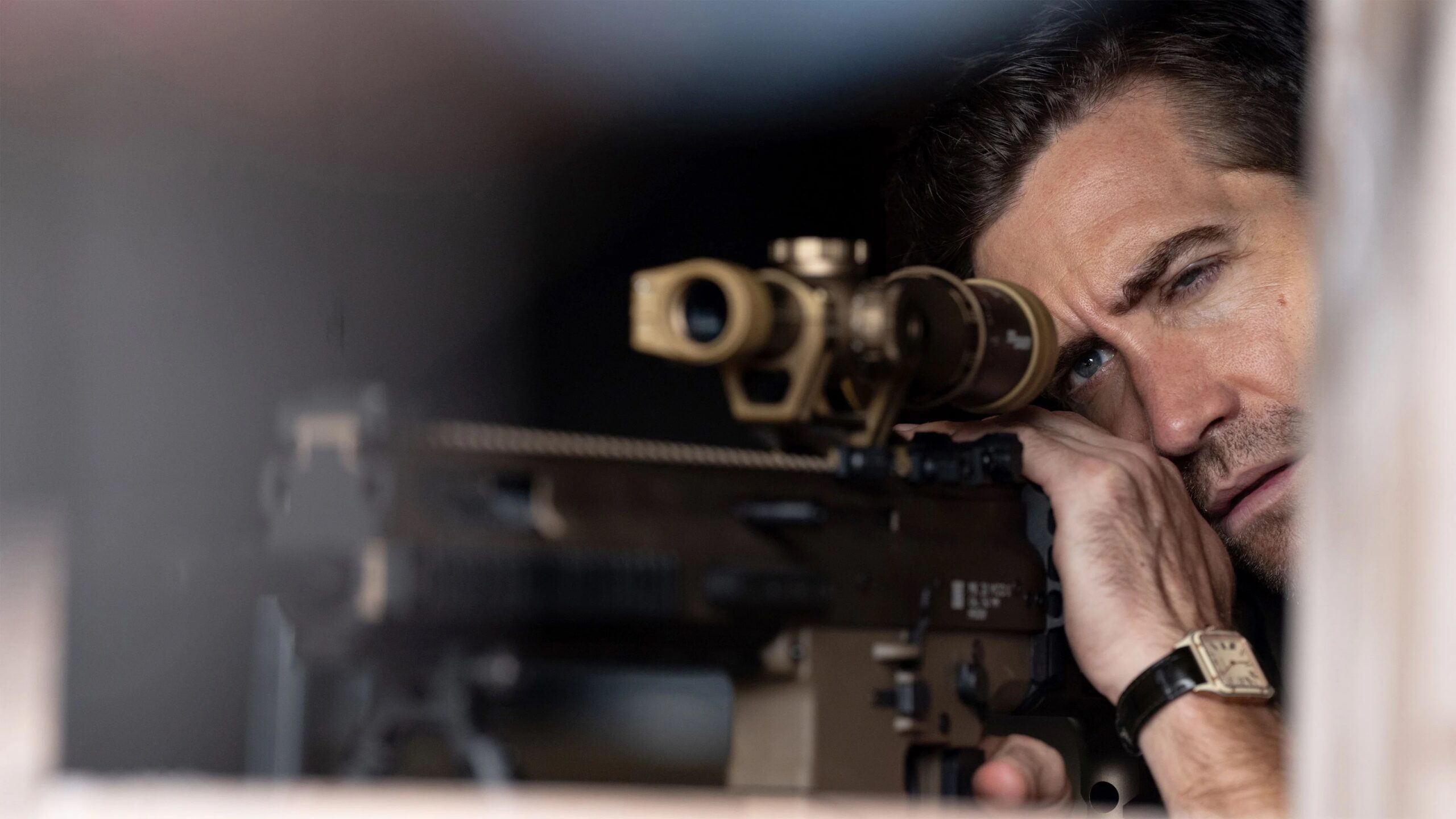 Jake Gyllenhaal als Bronco im Film In the Grey zielt konzentriert durch das Zielfernrohr eines sandfarbenen Gewehrs. Sein Auge liegt dicht am Scope, die Umgebung ist dunkel und unscharf, was die fokussierte, spannungsgeladene Stimmung verstärkt. Am Handgelenk trägt er eine rechteckige Uhr, während das goldene Glas des Visiers warm reflektiert.