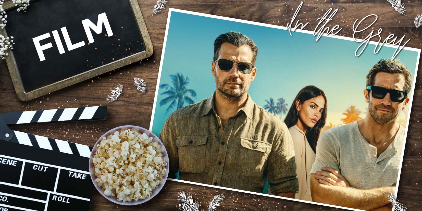 Filmstill zu In the Grey: Henry Cavill, Eiza González und Jake Gyllenhaal stehen gemeinsam vor Palmen im Sonnenlicht, alle mit Sonnenbrillen, während links im Bild eine Filmklappe, Popcorn und das Wort „Film“ auf einer Tafel liegen. Die Komposition verbindet Kino‑Ästhetik mit einem Promo‑Look der drei Hauptdarsteller.