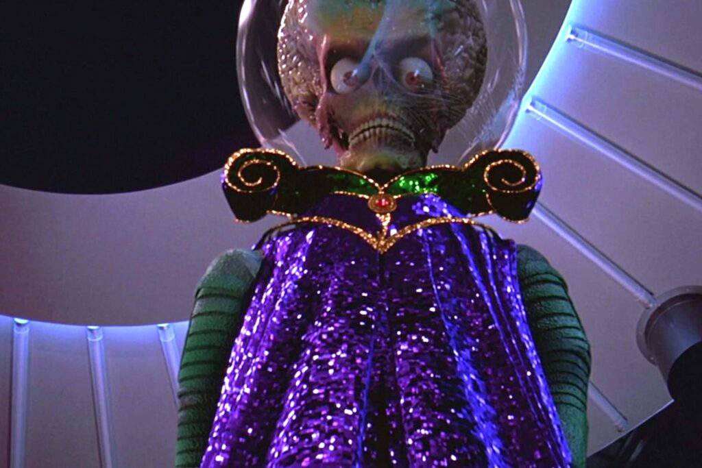 Ein Marsianer aus Mars Attacks! (1996) mit freiliegendem, großem Gehirn unter einer gläsernen Kuppel blickt bedrohlich nach vorn. Er trägt ein auffälliges, violett‑goldenes Gewand mit grünen Schulterstücken und steht in einer futuristischen Umgebung mit metallischen Strukturen und grellem Licht. | Die 5 kreativsten außerirdischen Wesen