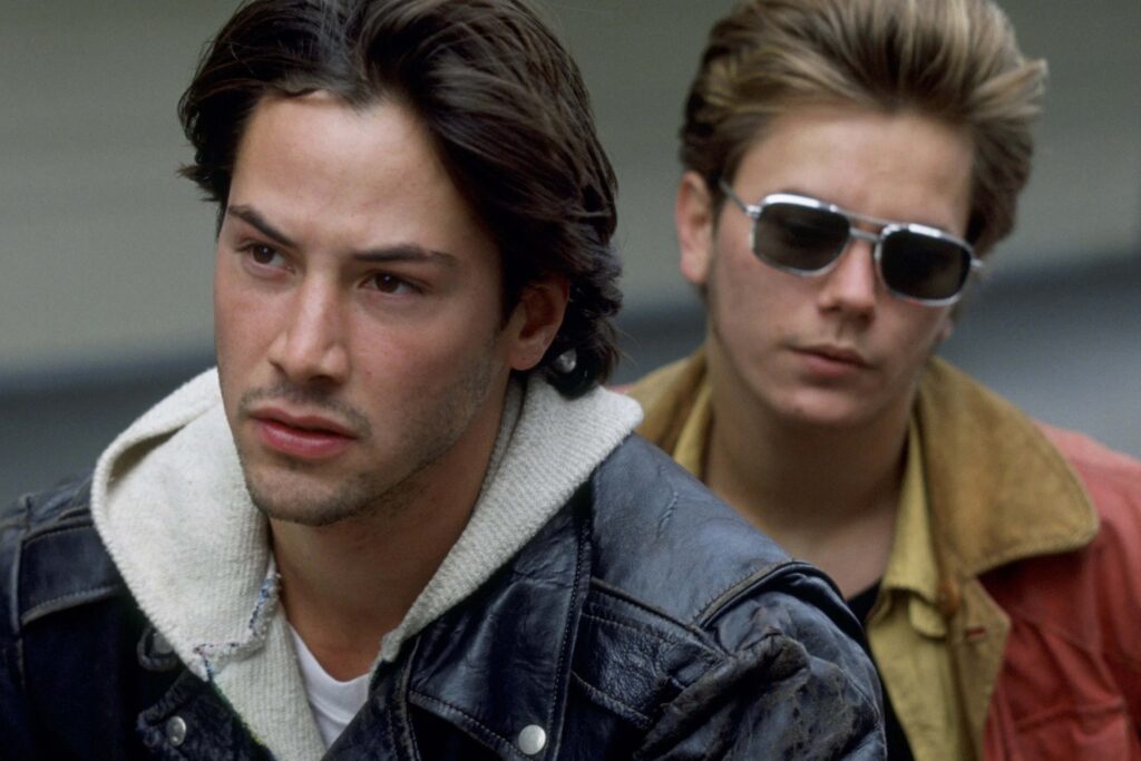 Keanu Reeves und River Phoenix in „My Private Idaho“ in einer Nahaufnahme im Freien: Reeves mit dunklem Haar, Lederjacke und konzentriertem Blick im Vordergrund, Phoenix mit Sonnenbrille und lässigem Outfit im Hintergrund. Die Szene betont die melancholische, suchende Stimmung des Films. | Media Monday #775