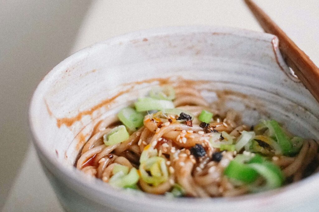 Schüssel mit würzigen Nudeln, garniert mit Frühlingszwiebeln, Sesam und Chili; Nahaufnahme eines frisch angerichteten asiatischen Nudelgerichts mit Holzstäbchen. | Media Monday #776