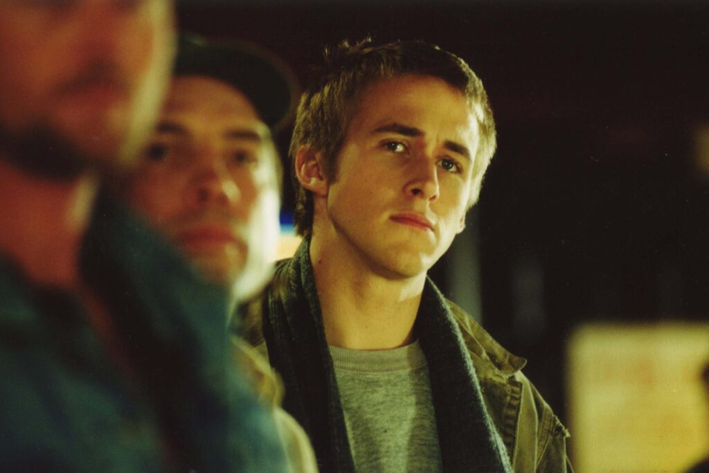 Ryan Gosling als Roy Chutney in The Slaughter Rule steht in einem dunklen Innenraum im Fokus, während zwei unscharfe Personen im Vordergrund vorbeigehen; sein Blick wirkt nachdenklich und distanziert. | Die 5 besten Coming-of-Age-Filme