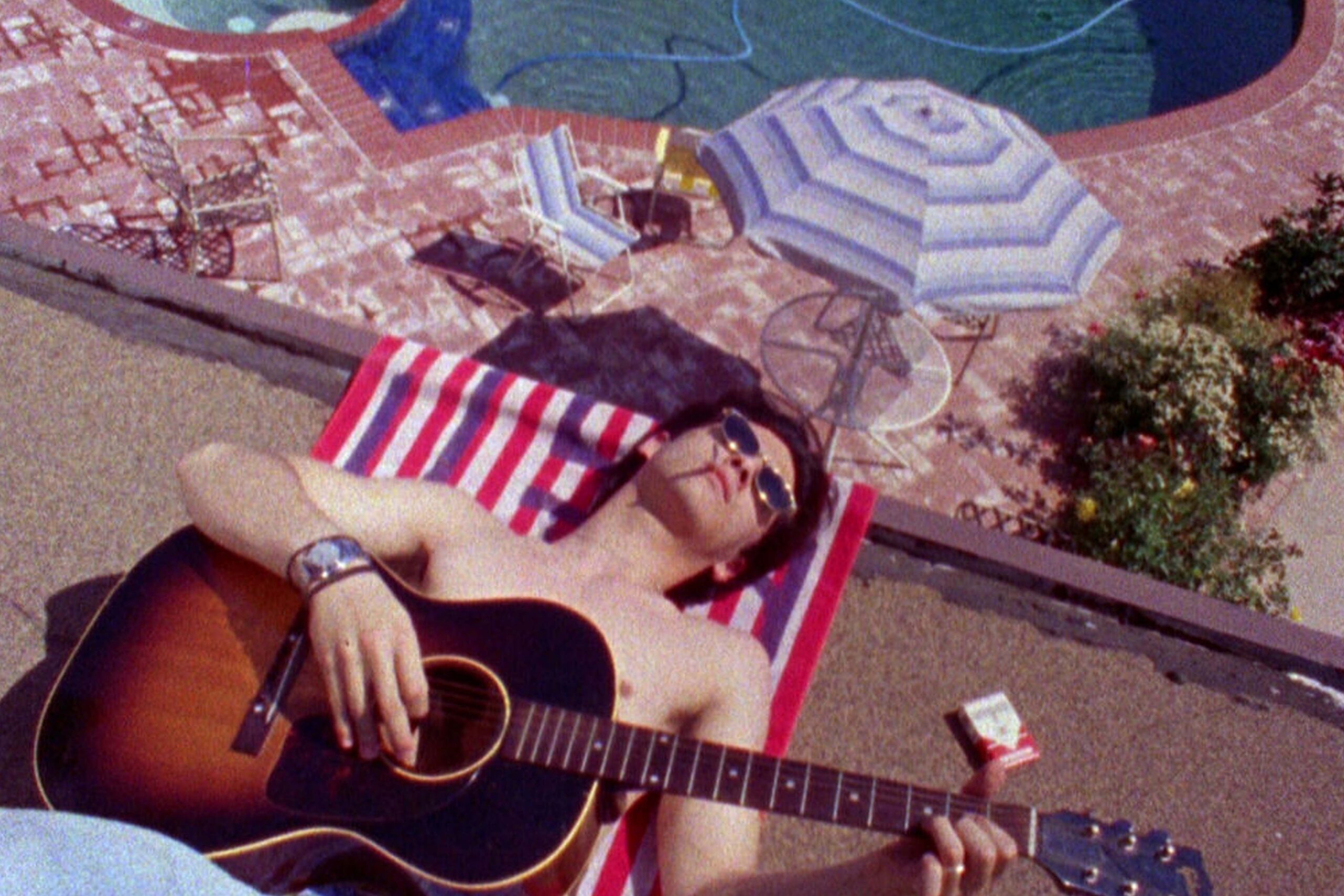 Andy (James Duval) aus Totally F***ed Up liegt entspannt auf einem gestreiften Handtuch am Pool, trägt Sonnenbrille und hält eine Akustikgitarre; Zigarettenpackung, Sonnenschirm und sommerliche Umgebung verstärken die lässige, melancholische Stimmung der Szene. | Die 5 besten Coming-of-Age-Filme
