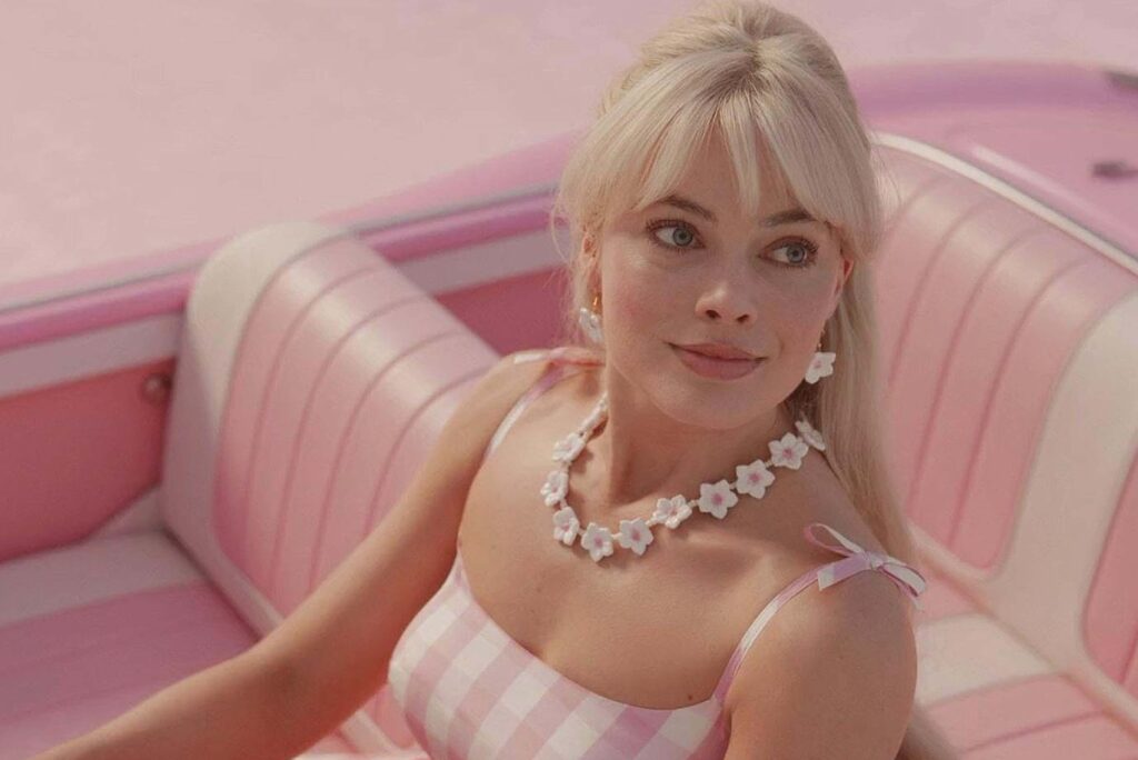 Margot Robbie sitzt als Barbie in einem pinken Cabrio mit pastellfarbenem Innenraum. Sie trĂ€gt ein rosaâweiĂ kariertes Kleid mit Schleifen an den TrĂ€gern, eine weiĂe Blumenhalskette und passende Ohrringe. Ihr blondes Haar ist im Pony gestylt. Die gesamte Szene ist in krĂ€ftigen Pinktönen gehalten und wirkt wie eine stilisierte, nostalgische Puppenwelt. | Media Monday #782