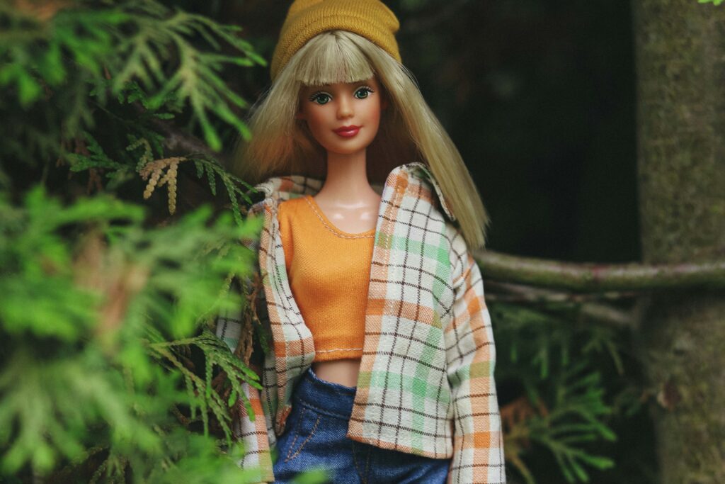 Eine blonde Barbie mit grünen Augen steht draußen zwischen dichtem Grün. Sie trägt eine gelbe Strickmütze, ein orangefarbenes Top, ein kariertes Hemd und Jeans. Die Szene wirkt natürlich, als würde die Puppe im Wald oder Garten posieren. | Media Monday #780