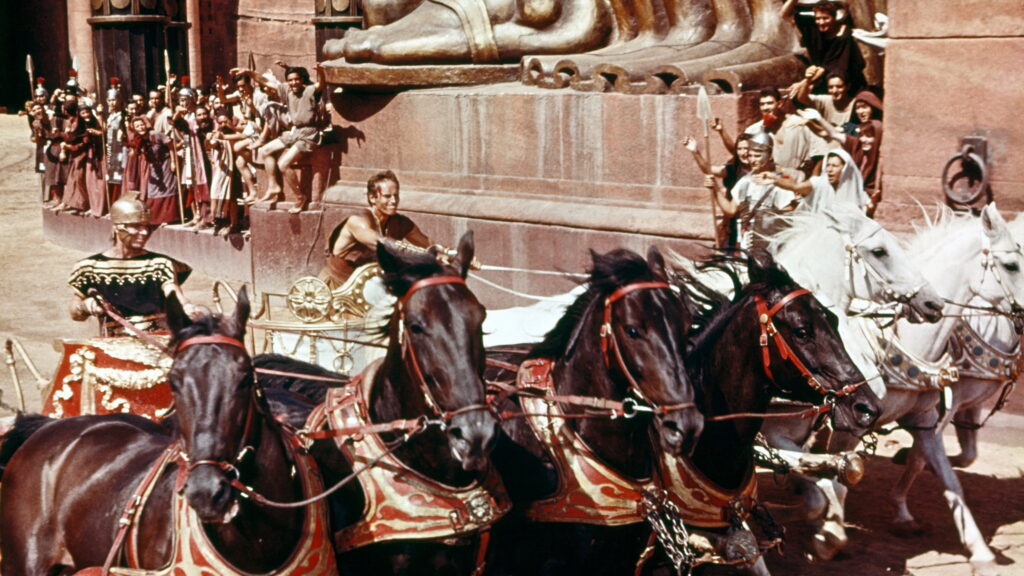 Ben Hur steht auf einem Wagen, der von 4 weißen Pferden gezogen wird. Links neben ihm ein anderer Wagen mit einem weiteren Fahrer.