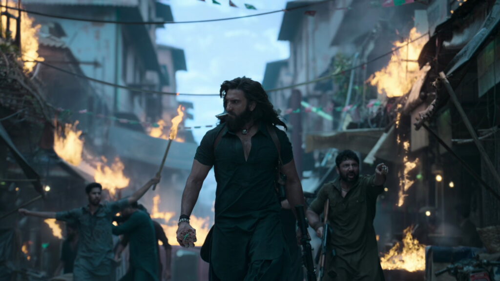 Das Bild zeigt Ranveer Singh mit einem Gewehr vor einer brennenden Häuserzeile in einer Szene des Films Dhurandhar: The Revenge. Top 5 Filme über 180 Minuten.