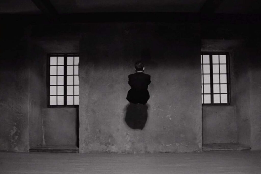 Eine Person steht gebeugt an einer Hauswand, zwischen zwei Fenstern. Das Bild wirkt bedrohlich, surreal und düster und gibt den Kern von Ingmar Bergmans "Die Stunde des Wolfs" wieder | Media Monday #780