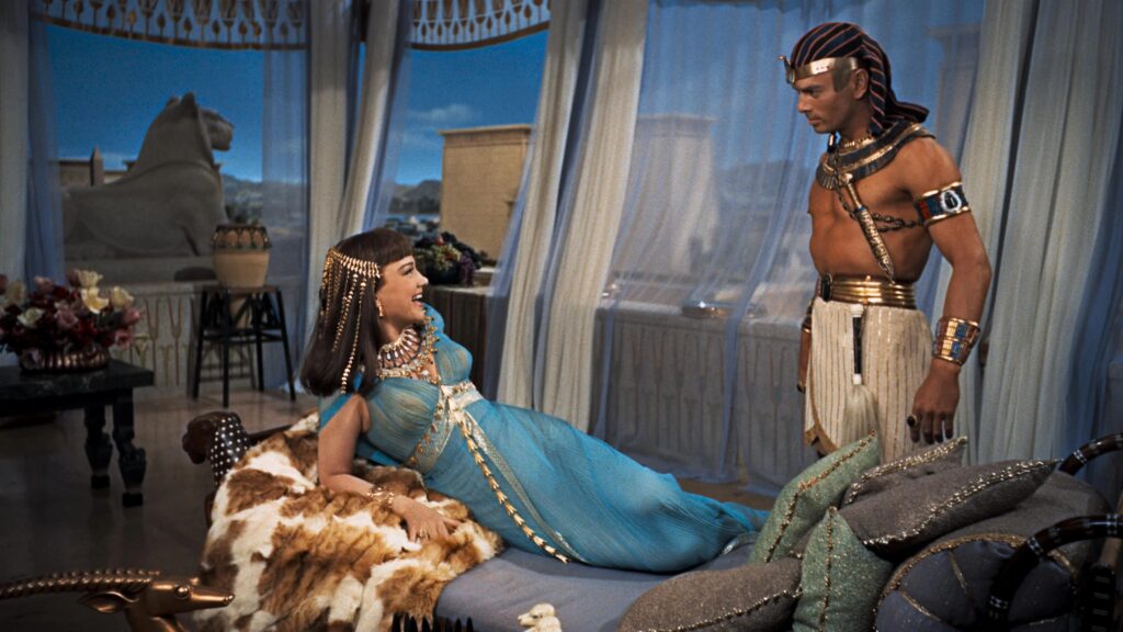Anne Baxter als Nefretiri und Yul Brynner als Pharao Ramses begegnen sich in „Die zehn Gebote“ (1956) in einer prachtvollen Gemachszene. Während Nefretiri in einem eleganten türkisfarbenen Gewand entspannt auf einer Liege ruht, steht Ramses in königlicher ägyptischer Kleidung vor ihr. Luxuriöse Vorhänge, kostbarer Schmuck und antike Dekorationen unterstreichen die opulente Ausstattung des Monumentalfilms. Top 5 Filme über 180 Minuten.