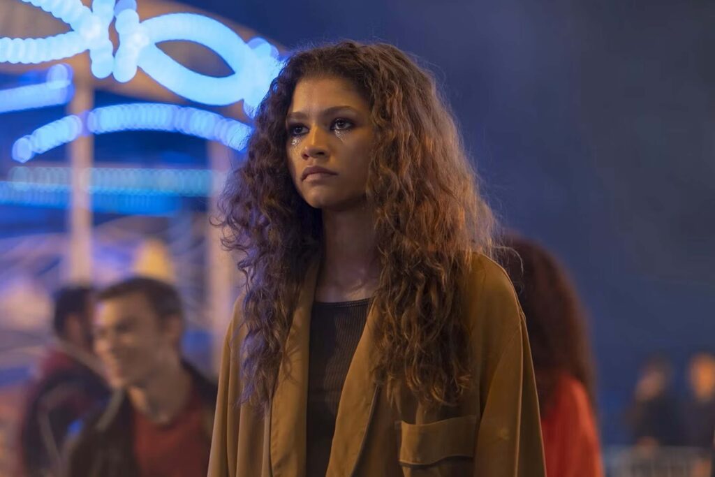 Zendaya als Rue Bennett steht nachts vor blau leuchtenden Neonlichtern. Sie trägt einen braunen Mantel über dunkler Kleidung, ihr Blick wirkt nachdenklich. Die Szene ist kontrastreich ausgeleuchtet, mit warmen Tönen im Vordergrund und kühlem Neonlicht im Hintergrund. | Media Monday #779