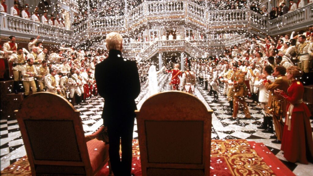 Das Bild zeigt einen Thronsaal während einer Vermählung. Es stammt aus dem Film Hamlet. Im Vordergrund ist die Titelfigur, gespielt von Kenneth Branagh, von hinten zu sehen. Top 5 Filme über 180 Minuten.