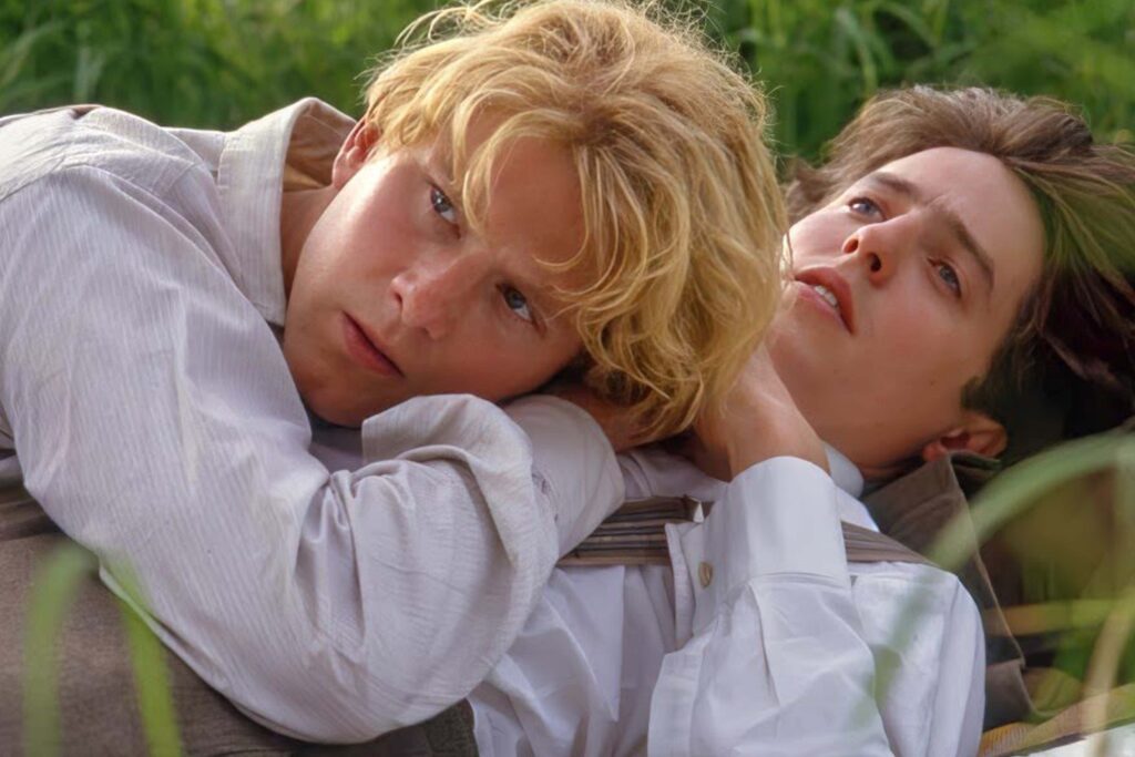 Maurice Hall (James Wilby) und Clive Durham (Hugh Grant) liegen eng beieinander im Gras einer Wiese; Maurice ruht mit dem Kopf auf Clives Brust, beide tragen helle Kleidung. Die Szene wirkt ruhig, zärtlich und zeigt ihre enge Verbindung in der Natur. | Media Monday #781