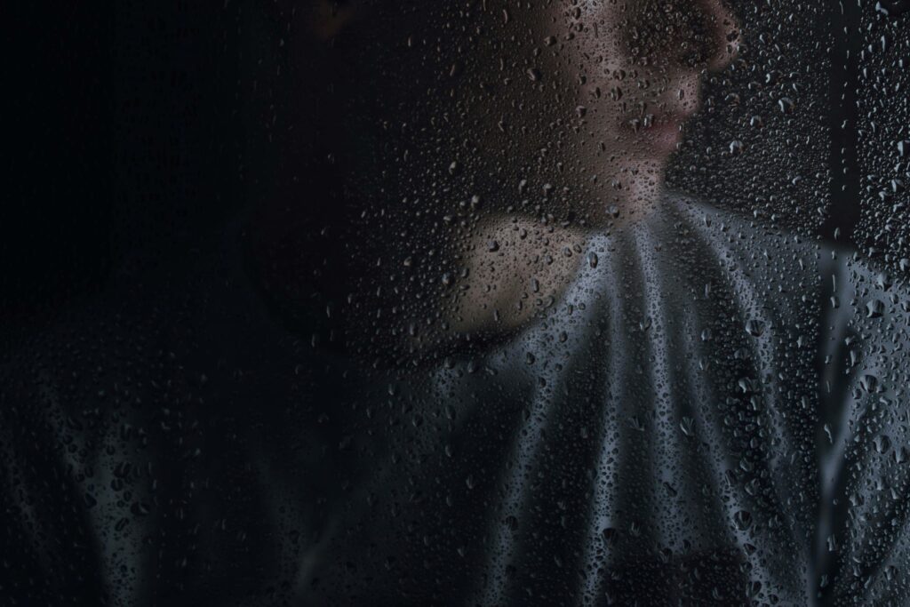 Verschwommenes Porträt einer Person hinter einer regennassen Glasscheibe; das Gesicht ist nur schemenhaft erkennbar, die Wassertropfen auf dem Glas stehen im Fokus und erzeugen eine ruhige, melancholische Stimmung. | Media Monday #781