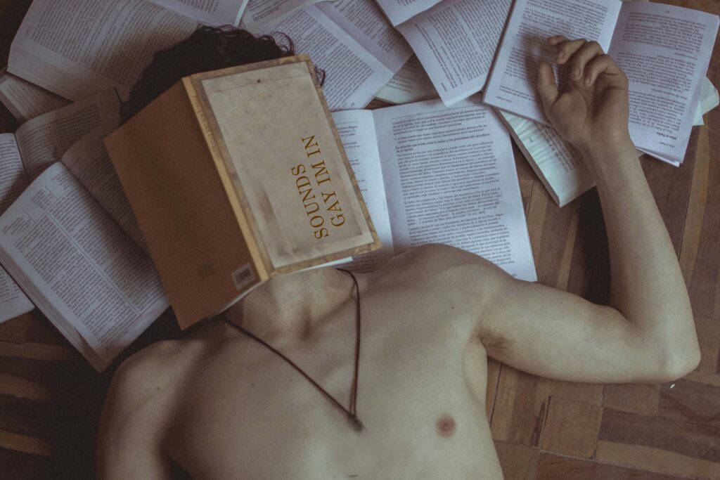 Oberkörper einer Person, die auf einem Holzboden zwischen offenen Büchern liegt; ein Buch mit der Aufschrift ‚SOUNDS GAY I’M IN‘ bedeckt das Gesicht. Die Szene wirkt verspielt, queer‑ästhetisch und leicht chaotisch, mit Fokus auf Büchern und Selbstironie. | Media Monday #781