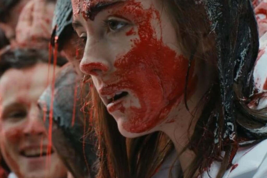 Justine (Garance Marillier) steht in der Ritualszene aus „Raw“ dicht von anderen Studierenden umgeben; ihr Gesicht, Haar und weißes Oberteil sind vollständig mit einer dicken roten Flüssigkeit überzogen, die wie Blut wirkt. Die Menschenmenge im Hintergrund ist ebenfalls bespritzt, wodurch die Szene intensiv, chaotisch und rituell aufgeladen erscheint. | Die 5 Besten Body Horror Film