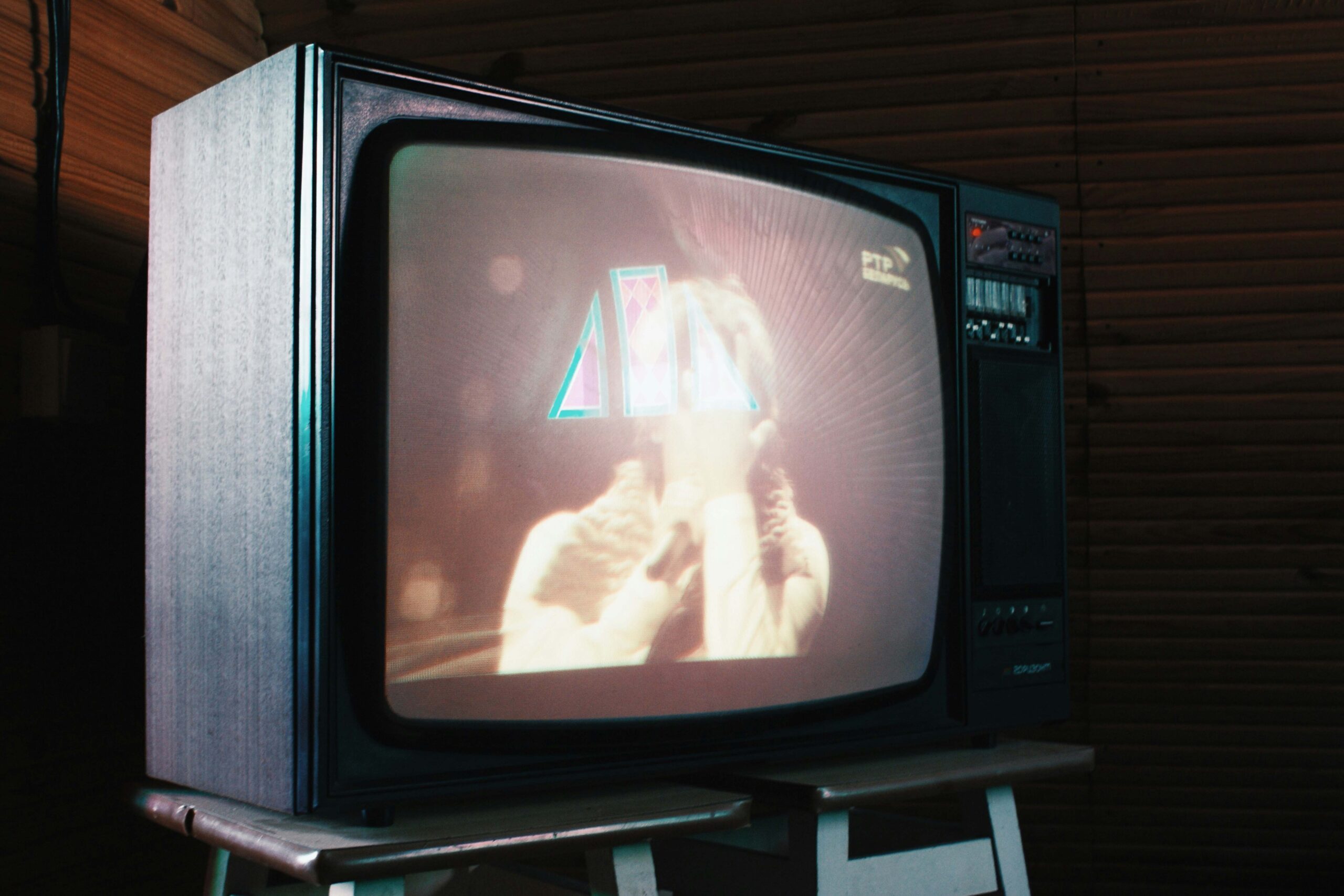 Alter Röhrenfernseher auf einem kleinen Holztisch, der eine unscharfe TV‑Sendung zeigt; warmes Retro‑Leuchten und Holzvertäfelung im Hintergrund erzeugen nostalgische Anmutung und erinnern an Zeiten, in denen Serien kontinuierlich im Fernsehen liefen. | Media Monday #794