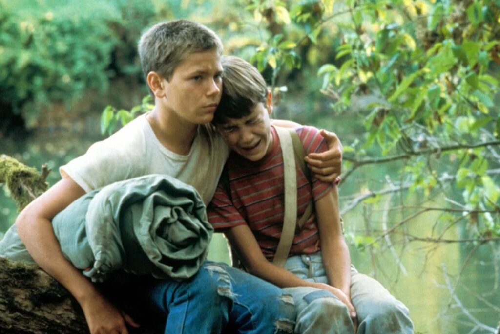 Chris Chambers (River Phoenix) tröstet den sichtlich aufgewühlten Gordie Lachance (Wil Wheaton), während die beiden Jungen am Ufer eines Waldflusses sitzen; die ruhige Naturkulisse und die innige Geste spiegeln die emotionale Tiefe und Freundschaft des Coming‑of‑Age‑Films „Stand by Me – Das Geheimnis eines Sommers“ wider. | Themenübersicht Juli 2026