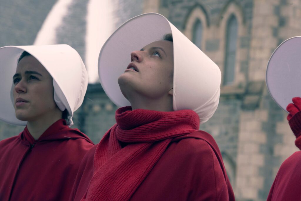 June Osborne steht in der Mitte, gekleidet in den typischen roten Habit und die weiße Haube der Mägde aus The Handmaid’s Tale: Der Report der Magd. Neben ihr stehen zwei weitere Mägde in gleicher Kleidung. Sie befinden sich vor einem steinernen Gebäude mit Rundbogenfenstern, und June blickt leicht nach oben, während die anderen nach vorn schauen. | Media Monday #780