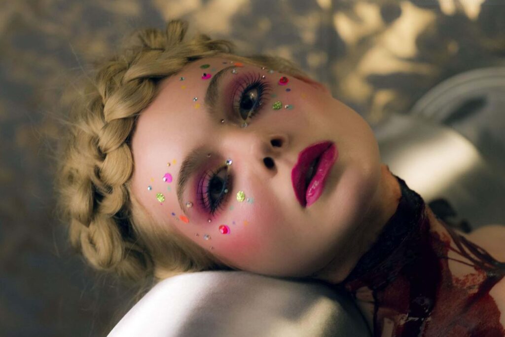 Elle Fanning liegt in „The Neon Demon“ auf einer glänzenden Oberfläche; ihr Gesicht ist mit farbigen Strasssteinen, Glitzer und intensivem pinkem Make‑up verziert, während hell geflochtenes Haar wie eine Krone arrangiert ist. Auf ihrem Hals und Brustbereich ist dunkle, blutähnliche Flüssigkeit zu sehen, was der Szene einen kontrastreich-düsteren, stilisierten Look verleiht. | Die 5 Besten Body Horror Film