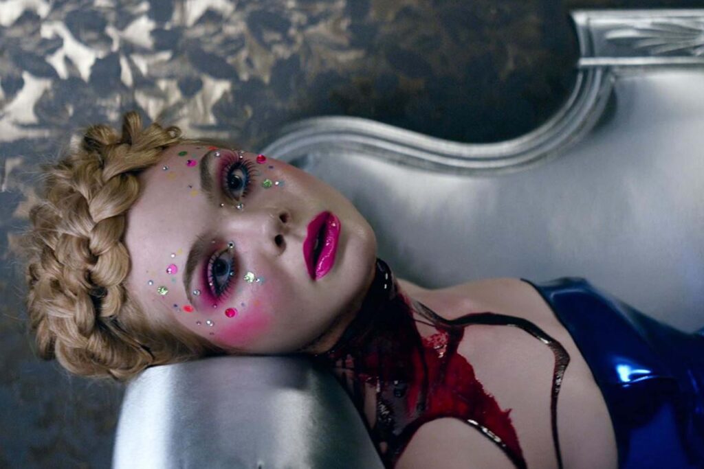 Elle Fanning als Jesse in „The Neon Demon“ liegt auf einer metallisch glänzenden Oberfläche; mit geflochtener Krone, intensiv pinkem Make‑up, glitzernden Strasssteinen im Gesicht, blau schimmerndem Outfit und schwarzem Spitzenchoker wirkt die Szene hyperstilisiert und kühl‑surreal, typisch für die ästhetische Bildsprache des Films. | Themenübersicht Juli 2026