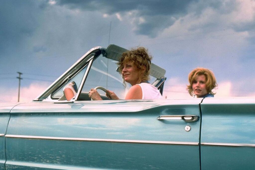 Thelma und Louise sitzen in einem hellblauen Cabrio mit offenem Verdeck. Thelma fährt, Louise sitzt daneben und blickt zu ihr herüber. Der Himmel ist leicht bewölkt, im Hintergrund steht ein Telefonmast. Die Szene vermittelt ein Gefühl von Freiheit, Aufbruch und gemeinsamer Reise. | Media Monday #779