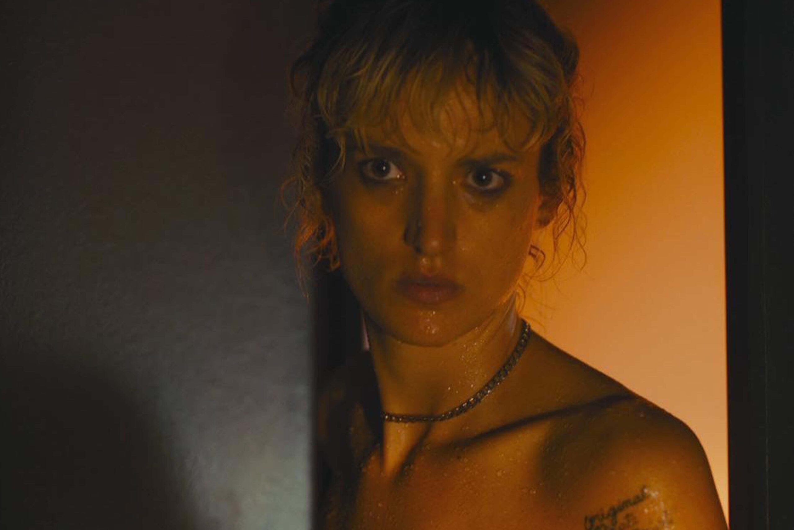 Alexia (Agathe Rousselle) steht in einer dunklen Umgebung, von warmem orangefarbenem Licht auf der einen und kühlerem Schatten auf der anderen Seite modelliert. Ihre Haut und Haare wirken feucht, ein dünner Kettenanhänger liegt auf der Brust, und am Oberarm ist ein Tattoo sichtbar. Die starke Licht‑Schatten‑Dramaturgie betont die körperliche Intensität und Identitätsauflösung, die „Titane“ prägt. | Die 5 Besten Body Horror Film