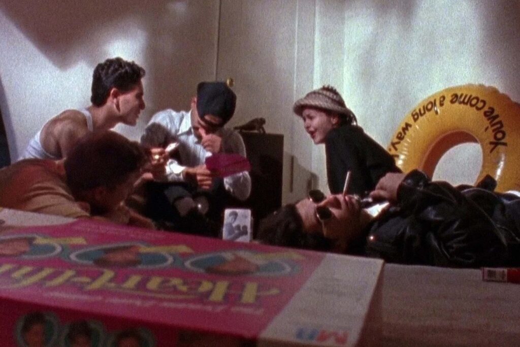 Szene aus „Totally F*ed Up“ (1993): Eine Gruppe Jugendlicher sitzt und liegt in einem chaotischen Zimmer auf dem Boden. Eine Person trägt eine Sonnenbrille und eine Lederjacke, eine andere einen Hut und lächelt in die Runde. Vor ihnen steht ein buntes Party‑Spiel, im Hintergrund hängt ein gelber aufblasbarer Ring. Die Atmosphäre wirkt roh, improvisiert und wie ein intimer Moment ihres Alltags. | Media Monday #779