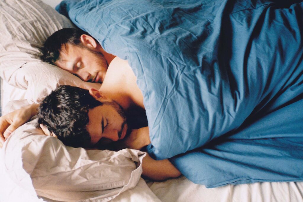 Szene aus „Weekend“ (2011): Russell (Tom Cullen) und Glen (Chris New) liegen eng beieinander im Bett und schlafen. Beide teilen sich eine blaue Decke, ihre Köpfe ruhen auf hellen Kissen. Das Licht ist weich und natürlich, die Stimmung intim, ruhig und voller Nähe. | Media Monday #779