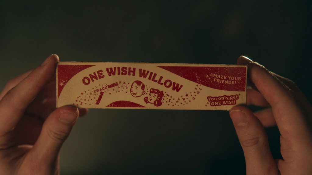 Zwei Hände halten ein schmales, längliches Schild im Vintage-Stil aus dem Film oder der Serie in die Kamera; das Design ist in Rot und Beige gehalten und zeigt den großen Schriftzug „ONE WISH WILLOW“. Neben den gezeichneten Gesichtern eines Jungen und eines Mädchens sind kleine Sterne sowie die Slogans „AMAZE YOUR FRIENDS!“ und „You only get ONE WISH“ aufgedruckt. Der Hintergrund ist düster und dunkelgrün schattiert, was eine geheimnisvolle, fast magische Stimmung erzeugt. © Universal Pictures Obsession