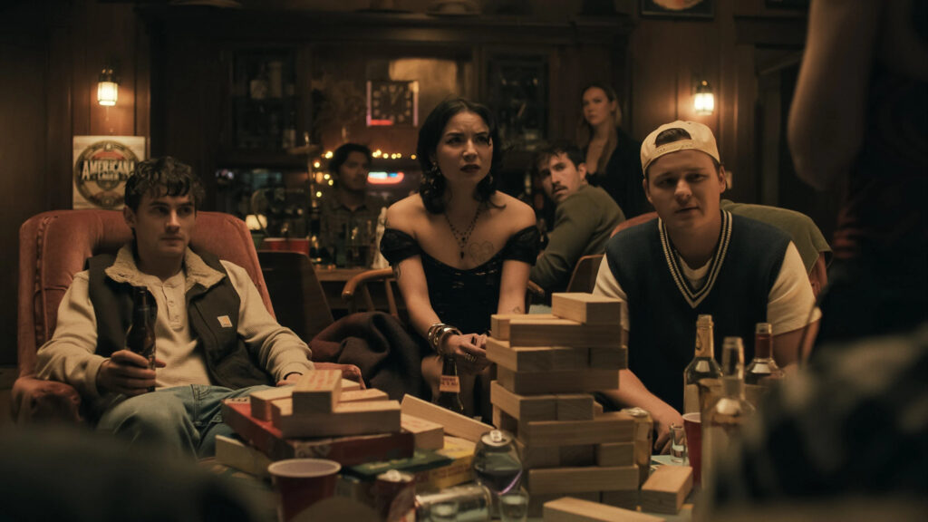 Die Schauspieler Michael Johnston (als Baron 'Bear' Bailey), Cooper Tomlinson (als Ian) und Megan Lawless (als Sarah Harper) sitzen gemeinsam auf einer Couch. Sie und die im Hintergrund sichtbaren Gäste blicken verstört in Richtung eines Charakters, der sich außerhalb des Sichtfelds vor der Kamera befindet. Vor der Gruppe auf dem Tisch stehen Alkoholflaschen und ein halb aufgebautes Jenga-Spiel. © Universal Pictures | Obsession 