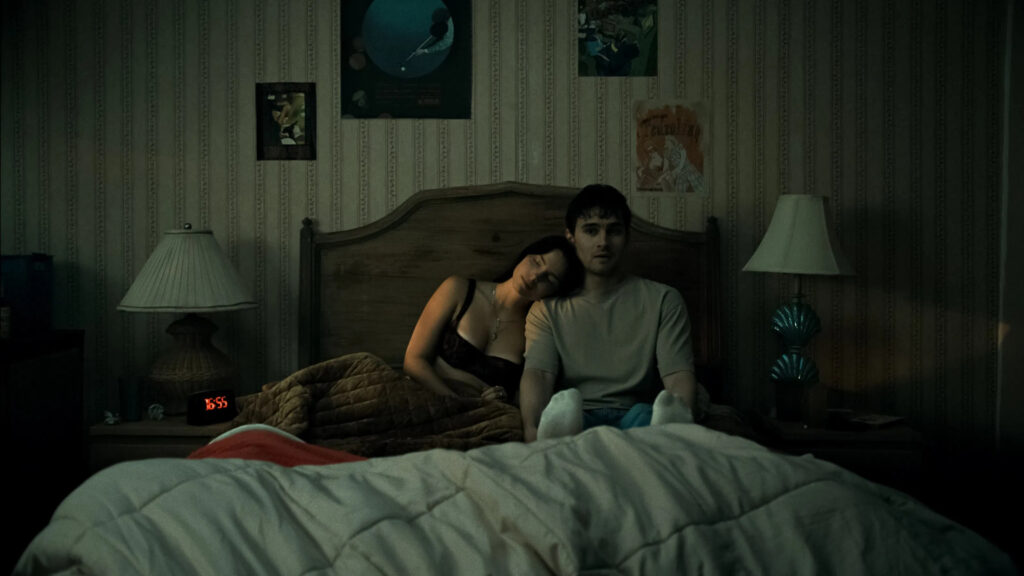 Der Schauspieler Michael Johnston (als Baron 'Bear' Bailey) und die Schauspielerin Inde Navarrette (als Niki Freemann) sitzen nebeneinander in einem Bett in einem schwach beleuchteten, rustikalen Schlafzimmer. Sie lehnt ihren Kopf an seine Schulter und hat die Augen geschlossen, während er mit einem nachdenklichen Blick nach vorne schaut. Links und rechts neben dem Bett steht jeweils eine Nachttischlampe, und an der gemusterten Tapete im Hintergrund hängen mehrere Poster. Im Vordergrund ist eine helle Bettdecke zu sehen. © Universal Pictures | Obsession 