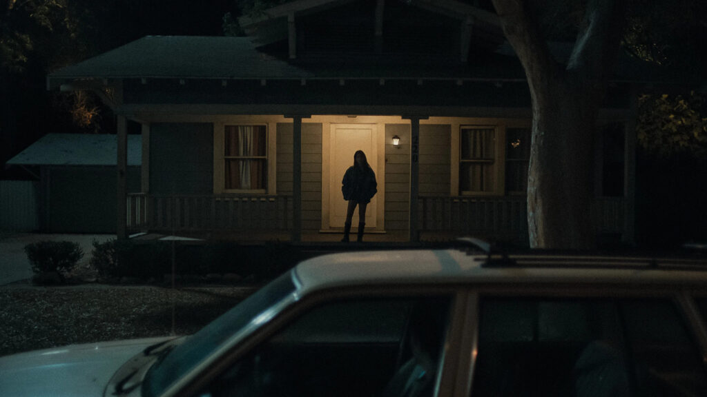 Die Schauspielerin Inde Navarrette (als Niki Freemann), die nachts allein auf der Veranda eines vorstädtischen Hauses steht. Sie ist eine Silhouette vor dem hellen Licht, und trägt eine dunkle Jacke und Stiefel. Das Haus ist ein einstöckiger Bungalow mit hellblau gestrichenen Holzpaneelen, einem kleinen Vordach und Fenstern, die ebenfalls von innen beleuchtet sind. Im Vordergrund ist die unscharfe Silhouette eines Autos mit Dachträger zu sehen, das auf der Straße geparkt ist. Die Szene ist dunkel und atmosphärisch, nur durch das Licht des Hauses beleuchtet. © Universal Pictures | Obsession 