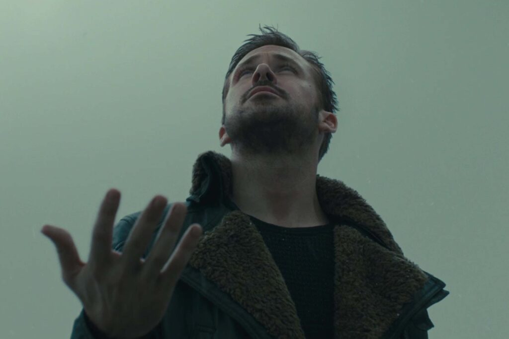 Ryan Gosling als Officer K steht in einem dunklen Mantel mit breitem Fellkragen unter einem grauen, nebligen Himmel. Er blickt nach oben und hält die Hand leicht geöffnet vor sich, um die ersten fallenden Schneeflocken zu fangen. Die Szene wirkt ruhig, melancholisch und typisch für die Atmosphäre von Blade Runner 2049. | Media Monday #787