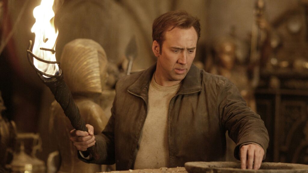 Benjamin Franklin Gates (Nicolas Cage) erkundet in „Das Vermächtnis der Tempelritter“ mit einer brennenden Fackel eine verborgene Schatzkammer. Umgeben von antiken Artefakten und einer ägyptischen Sarkophagfigur untersucht er aufmerksam ein steinernes Becken und folgt den nächsten Hinweisen auf der Suche nach dem legendären Schatz. | Die 5 besten Mystery-Abenteuerfilme