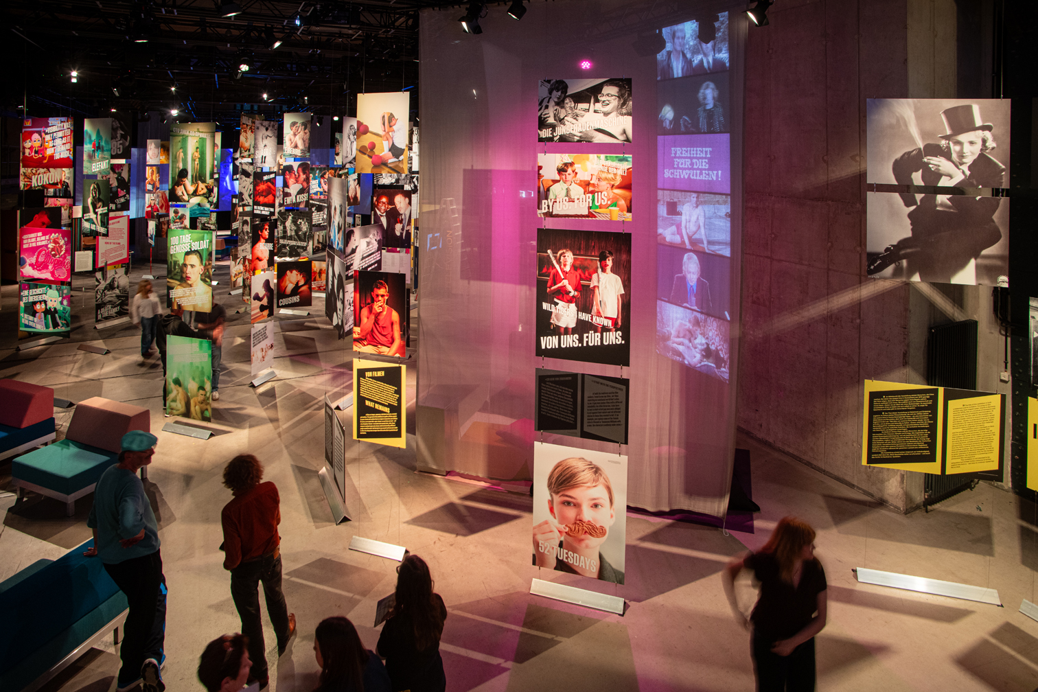 Foto der Ausstellung Inventing Queer Cinema in der Deutschen Kinemathek. Eine Halle mit vielen Bildern und Besucher:innen.