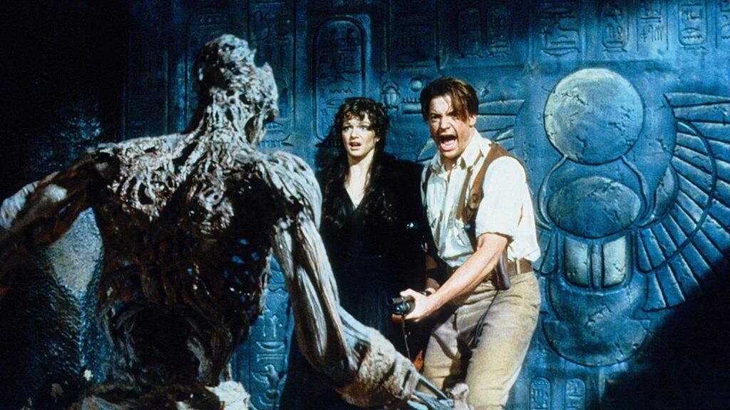 Rick O’Connell (Brendan Fraser) und Evelyn Carnahan (Rachel Weisz) stehen in „Die Mumie“ (1999) einer wiedererweckten Mumie in einer altägyptischen Grabkammer gegenüber. Vor den mit Hieroglyphen verzierten Wänden weicht das erschrockene Duo der bedrohlichen Kreatur zurück und fängt die Mischung aus Abenteuer, Horror und Spannung des Films ein. | Die 5 besten Mystery-Abenteuerfilme