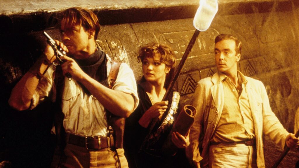 Rick O’Connell (Brendan Fraser), Evelyn Carnahan (Rachel Weisz) und Jonathan Carnahan (John Hannah) erkunden in „Die Mumie“ (1999) eine antike Grabkammer. Während Rick mit gezogener Pistole aufmerksam lauscht, hält Evelyn eine brennende Fackel und Jonathan eine Schriftrolle. Die mit Hieroglyphen bedeckten Wände und das warme Fackellicht verstärken die geheimnisvolle Abenteueratmosphäre des Films. | Die 5 besten Mystery-Abenteuerfilme