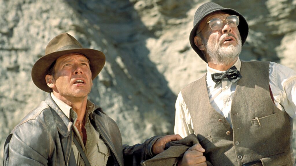 Indiana Jones (Harrison Ford) und sein Vater Professor Henry Jones Sr. (Sean Connery) blicken in „Indiana Jones und der letzte Kreuzzug“ aufmerksam nach oben. Vor einer hellen Felswand stehen die beiden Abenteurer Seite an Seite – Indiana mit Fedora und Lederjacke, sein Vater mit Brille, Fliege und Hut – und verkörpern die ikonische Vater-Sohn-Dynamik des Films. | Die 5 besten Mystery-Abenteuerfilme
