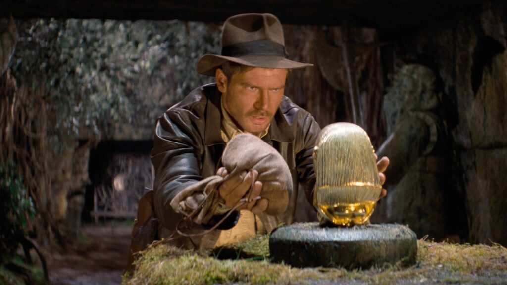 Indiana Jones (Harrison Ford) versucht in „Jäger des verlorenen Schatzes“ (1981) vorsichtig ein goldenes Götzenbild von seinem steinernen Sockel zu nehmen. Mit einem Sandsack in der Hand versucht der Archäologe das Gewicht auszugleichen, während die düstere Tempelkammer den ikonischen Auftakt des Abenteuerklassikers bildet.