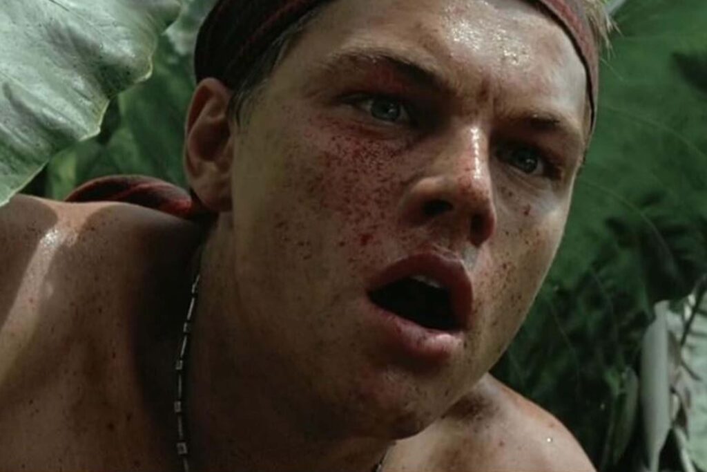 Nahaufnahme von Richard (Leonardo DiCaprio) aus The Beach (2000). Sein Gesicht ist blutverschmiert, der Mund leicht geöffnet, und er trägt ein Stirnband sowie eine dünne Kette. Er befindet sich zwischen großen grünen Blättern in einer dichten Dschungelumgebung, die eine angespannte und erschöpfte Stimmung vermittelt. | Themenübersicht August 2026