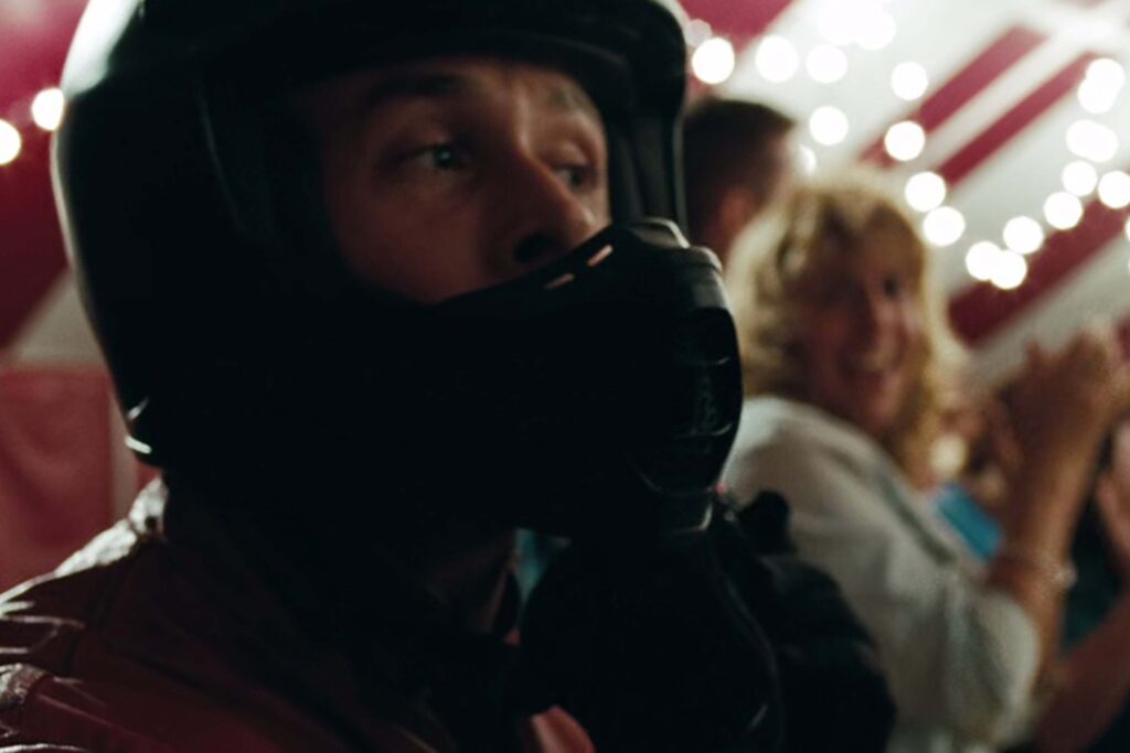 Ryan Gosling als Luke Glanton steht in einem schwarzen Motorradhelm und einer roten Jacke im Vordergrund. Hinter ihm applaudieren und lächeln mehrere Menschen unter einem Zelt mit Lichterkette und rot‑weißem Stoff, was der Szene einen festlichen, lebendigen Charakter verleiht. | Media Monday #787