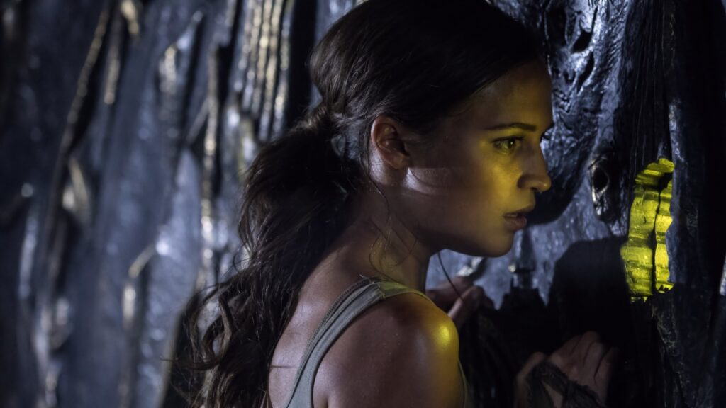Lara Croft (Alicia Vikander) untersucht in „Tomb Raider“ (2018) eine geheimnisvolle Wand mit einem leuchtenden Mechanismus. In der dunklen Grabkammer sucht sie konzentriert nach einem verborgenen Hinweis, während das schwache Licht die rätselhafte und abenteuerliche Atmosphäre der Szene betont.