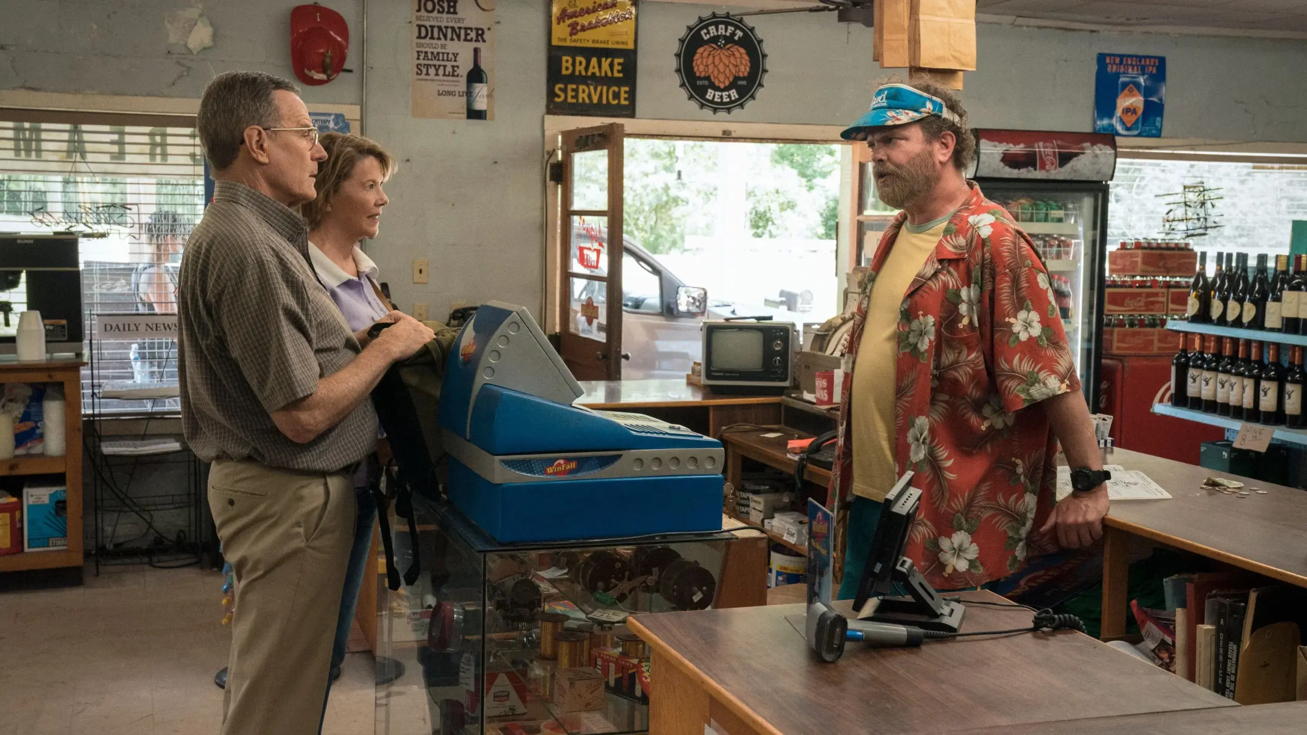 Jerry Selbee (Bryan Cranston) und Marge Selbee (Annette Bening) sprechen in „Jerry und Marge – Die Lottoprofis“ (2022) mit einem Ladenbesitzer hinter der Kasse eines kleinen Geschäfts. Während Jerry aufmerksam zuhört und Marge freundlich neben ihm steht, lehnt sich der Verkäufer in bunter Hawaii-Hemd-Kleidung über den Tresen. Die schlichte Einrichtung des Ladens mit Getränkeregalen, Werbeschildern und Kasse unterstreicht den bodenständigen Alltagsschauplatz der Komödie. | FYP Filme
