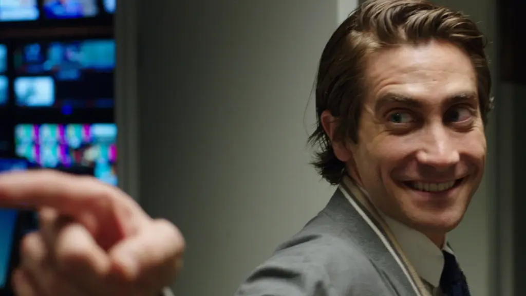 Lou Bloom (Jake Gyllenhaal) blickt in „Nightcrawler – Jede Nacht hat ihren Preis“ (2014) mit einem übertriebenen, beunruhigenden Grinsen zur Seite. Seine Haare sind streng nach hinten gekämmt, und er trägt eine graue Jacke über einem Hemd mit Krawatte. Im Vordergrund deutet eine unscharfe Hand mit dem Zeigefinger auf ihn, während im dunklen Hintergrund mehrere Monitore eines News-Studios aufleuchten. Die Nahaufnahme unterstreicht die manipulative und soziopathische Ausstrahlung des Protagonisten im modernen Medien-Thriller. | FYP Filme