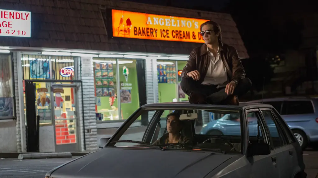 Lou Bloom (Jake Gyllenhaal) sitzt im Schneider- und Kontrollsitz auf dem Dach eines parkenden Autos vor einem beleuchteten Geschäft namens „Angelino's Bakery Ice Cream & Deli“. Er trägt eine Sonnenbrille, eine braune Lederjacke und blickt aufmerksam in die Nacht. Im Fahrzeuginneren sitzt sein Assistent Rick (Riz Ahmed) hinter dem Steuer und schaut durch die Windschutzscheibe. Die nächtliche Szene vor dem erleuchteten Laden unterstreicht das rastlose Lauern auf den nächsten Sensationseinsatz im Neo-Noir-Thriller. | FYP Filme